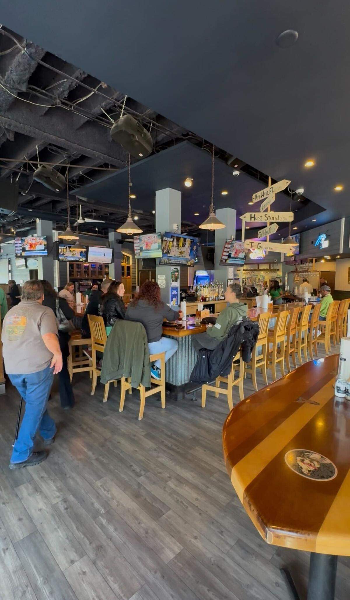 A wide shot of a bustling indoor bar and restaurant area. Several patrons are seated on light wood barstools at a long, wrap-around bar with a corrugated metal base. Multiple flat-screen TVs hang from the dark ceiling, showing sports. A decorative wooden signpost with directional arrows (pointing to “Key West, FL” and “Host Stand”) stands near the center. The flooring is a grey wood-plank style, and the space is lit by warm pendant lights and recessed ceiling lights.