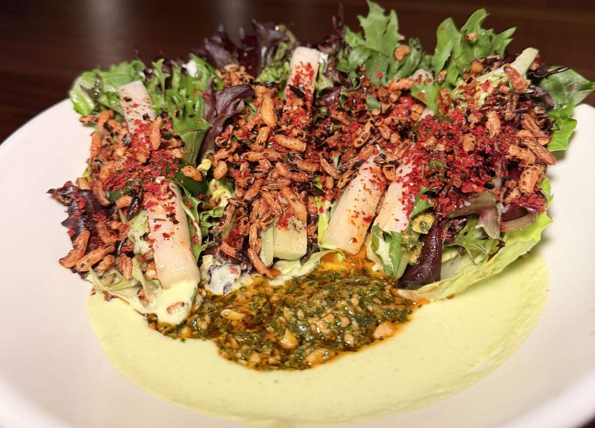 A vibrant, textured salad is served in a wide white bowl, featuring a mix of fresh greens and bold, colorful toppings. Interspersed among the greens are several vertical stalks of daikon The salad is heavily topped with a crunchy puffed rice or toasted grain garnish, accented with a bright red spice—likely sumac or beet powder.
