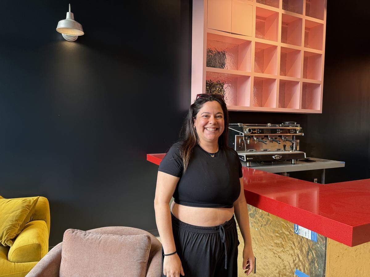 A bar owner smiling in a stylish space featuring a red bar, pink shelving, and colorful velvet furniture against a black wall.