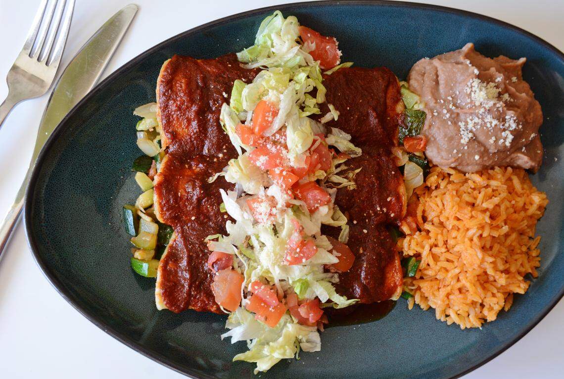 A plate of food, with two enchiladas covered in a dark red sauce and topped with shredded lettuce, chopped tomatoes, and grated white cheese. The enchiladas sit on a bed of sautéed zucchini. On the side are a scoop of refried beans topped with cheese and a pile of Spanish rice. A fork and knife are visible to the left of the plate.