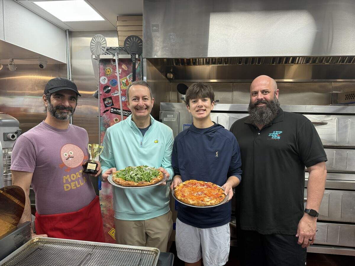 Four smiling people are standing side-by-side in a commercial kitchen in front of a large stainless steel pizza oven. The person on the far left wears a purple graphic t-shirt, a red apron, and a cap, and is holding a small gold trophy. The second person wears a light blue pullover and holds a metal tray with a baked pizza generously topped with fresh green arugula. The third person wears a dark blue hoodie and holds a tray with a classic cheese and tomato sauce pizza. The person on the far right wears a black polo shirt with the “Zepeddies Pizzeria” logo.