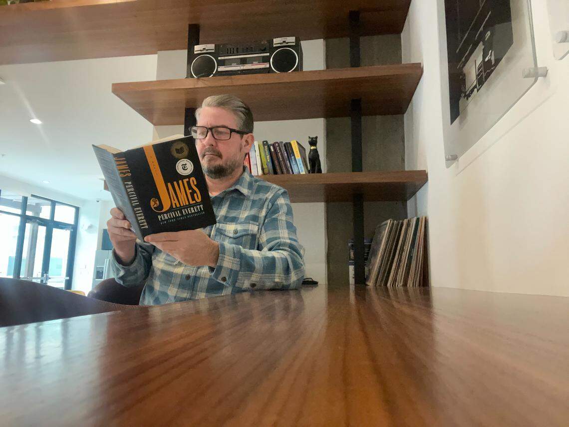 A person with glasses and a plaid shirt sits at a large wooden table, reading the novel “James” by Percival Everett. In the background are shelves holding a boombox, books, a black cat statue, and a stack of vinyl records.