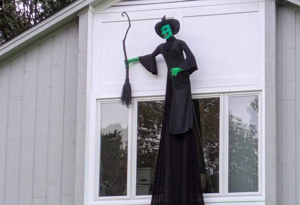 A wide shot of a large Halloween decoration of a witch mounted on the side of a house. The witch has green skin, a wide smile, a tall pointed black hat, and a long black dress, and is holding a black broomstick. It is positioned above a large window on the home’s light gray siding.