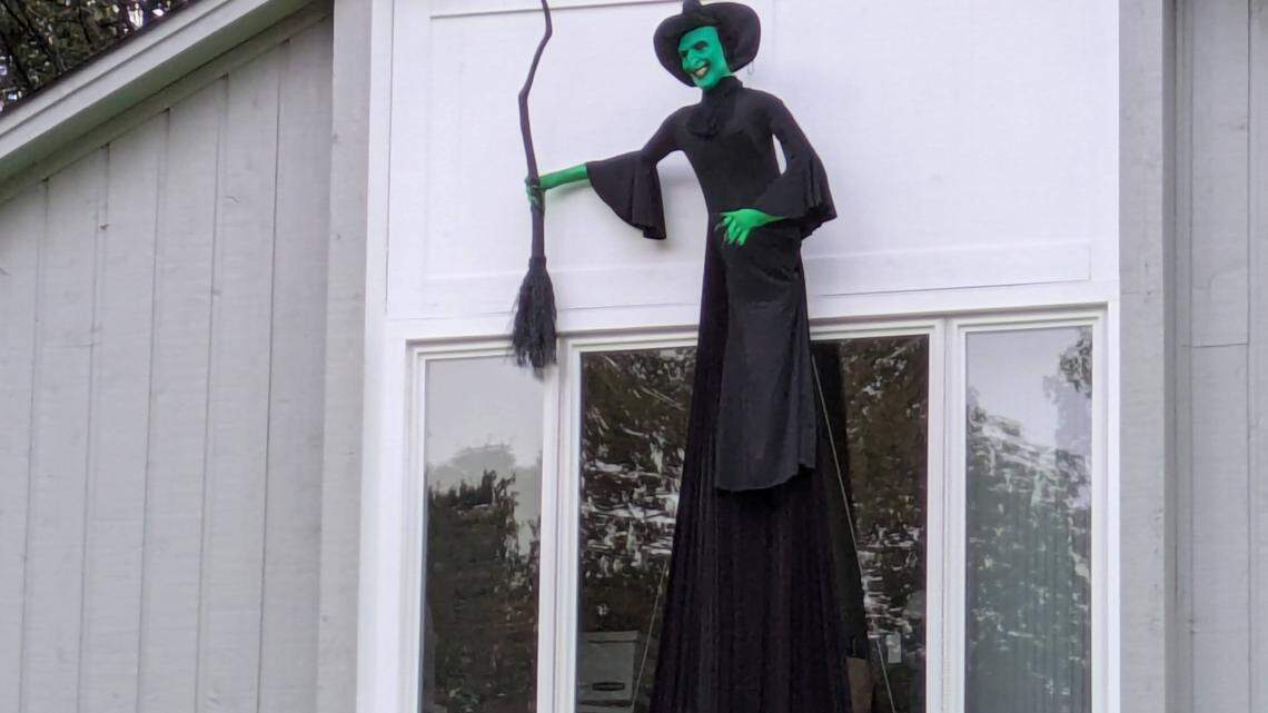 This two-story smiling Elphaba is the newest addition to the Halloween Trail at the South Charlotte home of Mark Barden and his wife. A wide shot of a large Halloween decoration of a witch mounted on the side of a house. The witch has green skin, a wide smile, a tall pointed black hat, and a long black dress, and is holding a black broomstick. It is positioned above a large window on the home’s light gray siding.