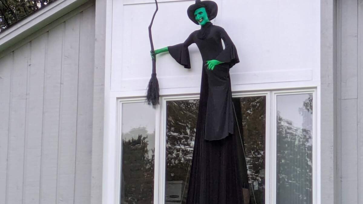 A wide shot of a large Halloween decoration of a witch mounted on the side of a house. The witch has green skin, a wide smile, a tall pointed black hat, and a long black dress, and is holding a black broomstick. It is positioned above a large window on the home’s light gray siding.