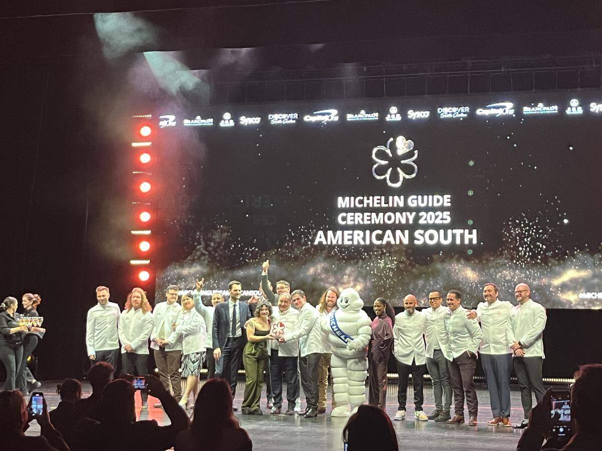A wide shot from the audience of the finale of the “MICHELIN GUIDE CEREMONY 2025 AMERICAN SOUTH.” A large group of chefs, mostly in white jackets, are gathered on stage, smiling and celebrating alongside the Michelin Man mascot. A large screen behind them displays the event’s title, and the stage is brightly lit.