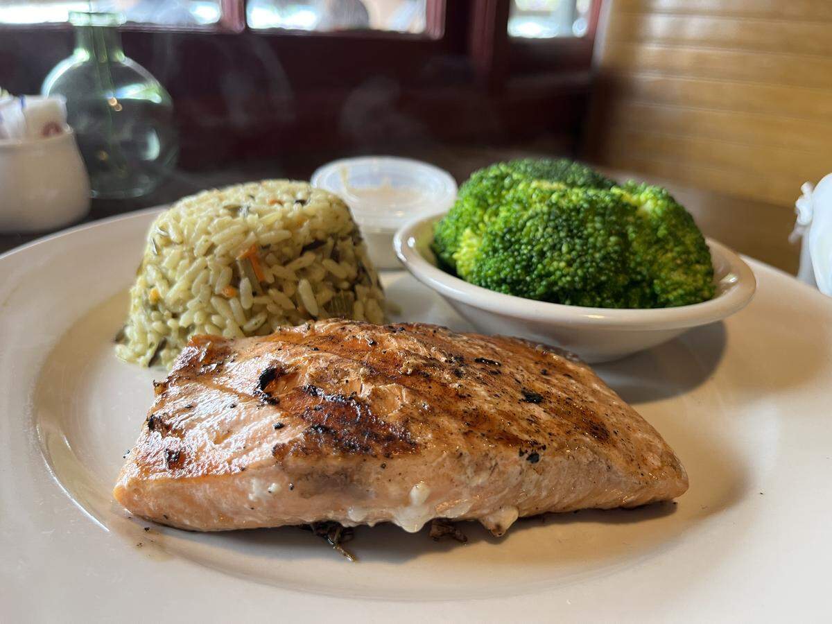 A hot meal served on an off-white plate. A large piece of grilled salmon with char marks is in the foreground. Behind it is a neatly rounded scoop of wild rice and a small bowl containing steamed broccoli florets. A small cup of white sauce is also on the plate.