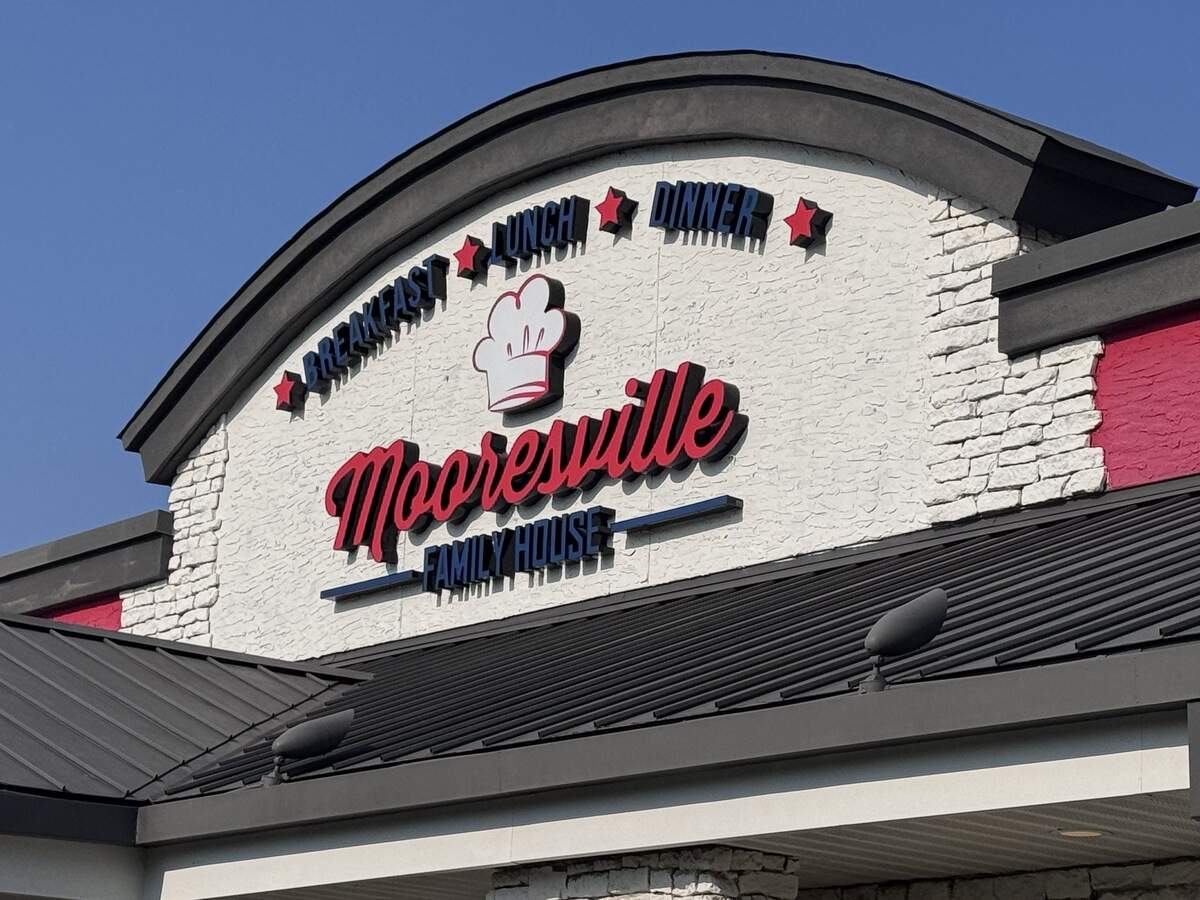 The exterior signage for “Mooresville Family House” restaurant set against a white faux-brick wall under a clear blue sky. A chef’s hat graphic sits above red script lettering, framed by black and blue text reading “Breakfast, Lunch, Dinner” separated by red stars.