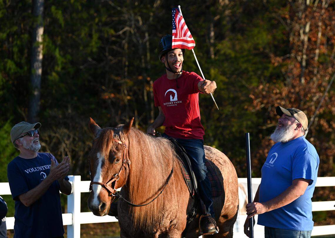 Chancellor Lee Adams, center, smiles as he pulls the flag from a holder while riding Roanie, a 22-year-old American quarter horse at Victory Farm on Friday, November 8, 2024. Looking on are volunteers Scott Hrvatin, left and Kelly Cheshire, right. Victory Farm is a therapeutic riding center in Gastonia.
