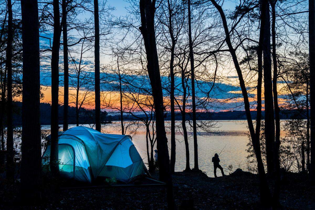 A glowing blue tent at a campsite along the wooded shore of a lake during a vibrant sunset, with a person silhouetted fishing from the water’s edge.