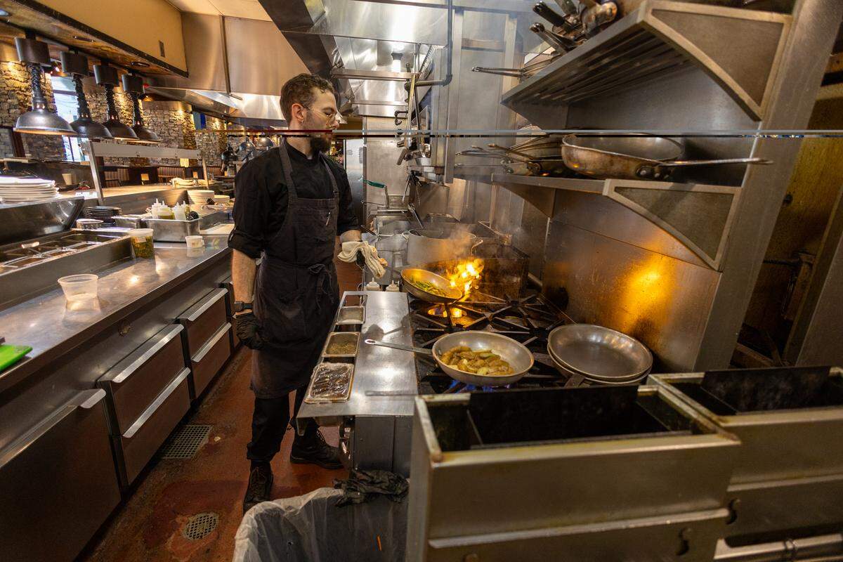 A wide shot of a professional restaurant kitchen. A chef wearing a black uniform and apron is focused on sautéing vegetables in a pan over an open gas flame. Stainless steel counters and industrial shelving filled with pans are visible.