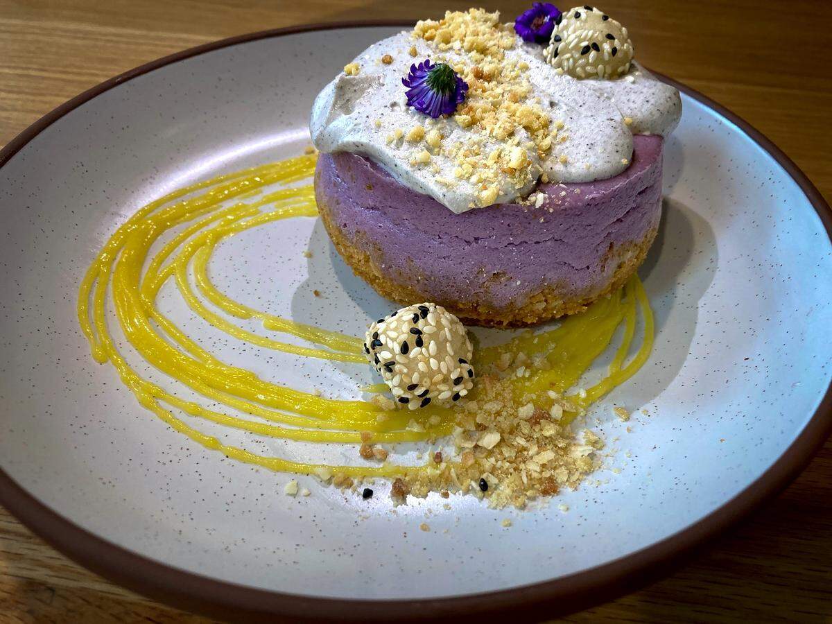 An elegant dessert of a mini ube cheesecake on a modern plate. The cheesecake has a crumb crust and a pale purple body, and is finished with a light grey mousse, toasted crumble, two small purple flowers, and several sesame-covered spheres. The plate is decorated with a looping drizzle of bright yellow sauce.
