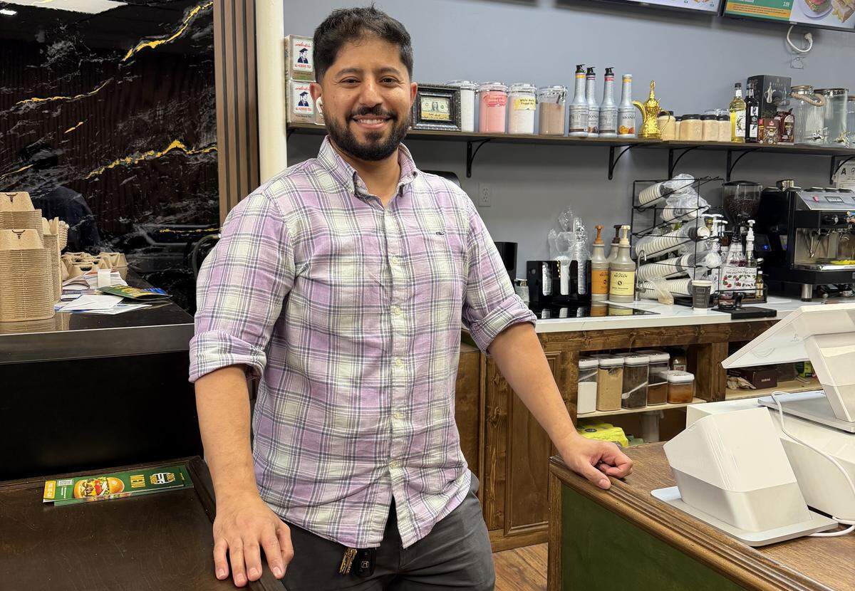 A medium shot of a smiling person with a short beard and a purple plaid shirt, standing behind a wooden service counter in a cafe. The background features a well-stocked beverage station with syrup bottles, a coffee machine, stacked cups, and shelving filled with jars and canisters.
