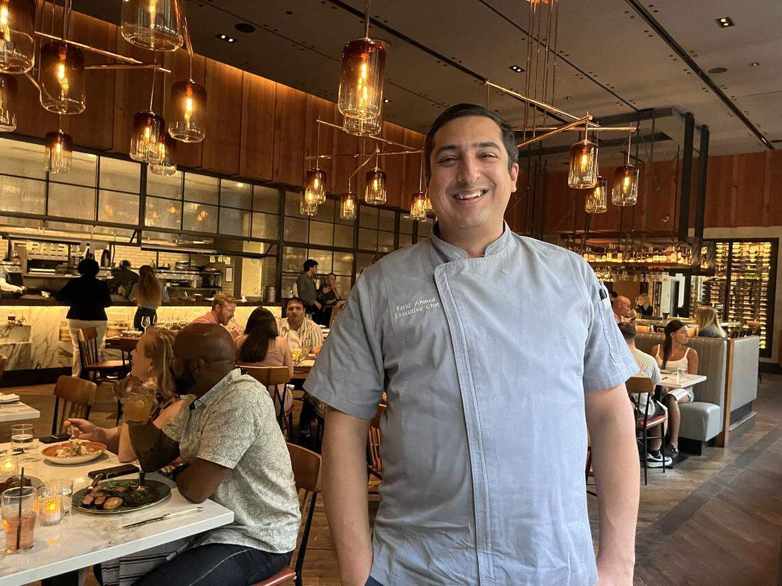 A portrait of a smiling chef in a light gray chef’s coat is standing in the middle of a bustling restaurant. The background is a large, modern dining room with other patrons seated at tables, and many hanging light fixtures are visible, creating a warm glow.