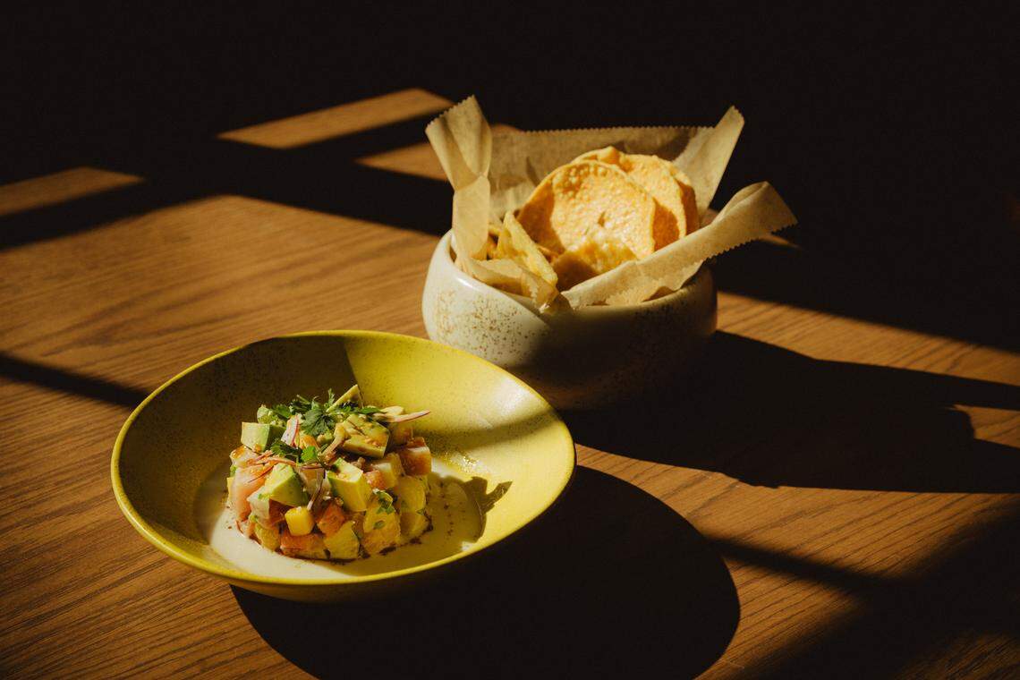 A high-angle shot of Ceviche Tropicoso presented in a bright yellow bowl. The ceviche is neatly molded into a circular stack, featuring visible chunks of white fish, bright green avocado, and mango. It sits in a pool of creamy white leche de tigre. In the background, a rustic ceramic bowl filled with golden-brown tortilla chips sits tucked in parchment paper.