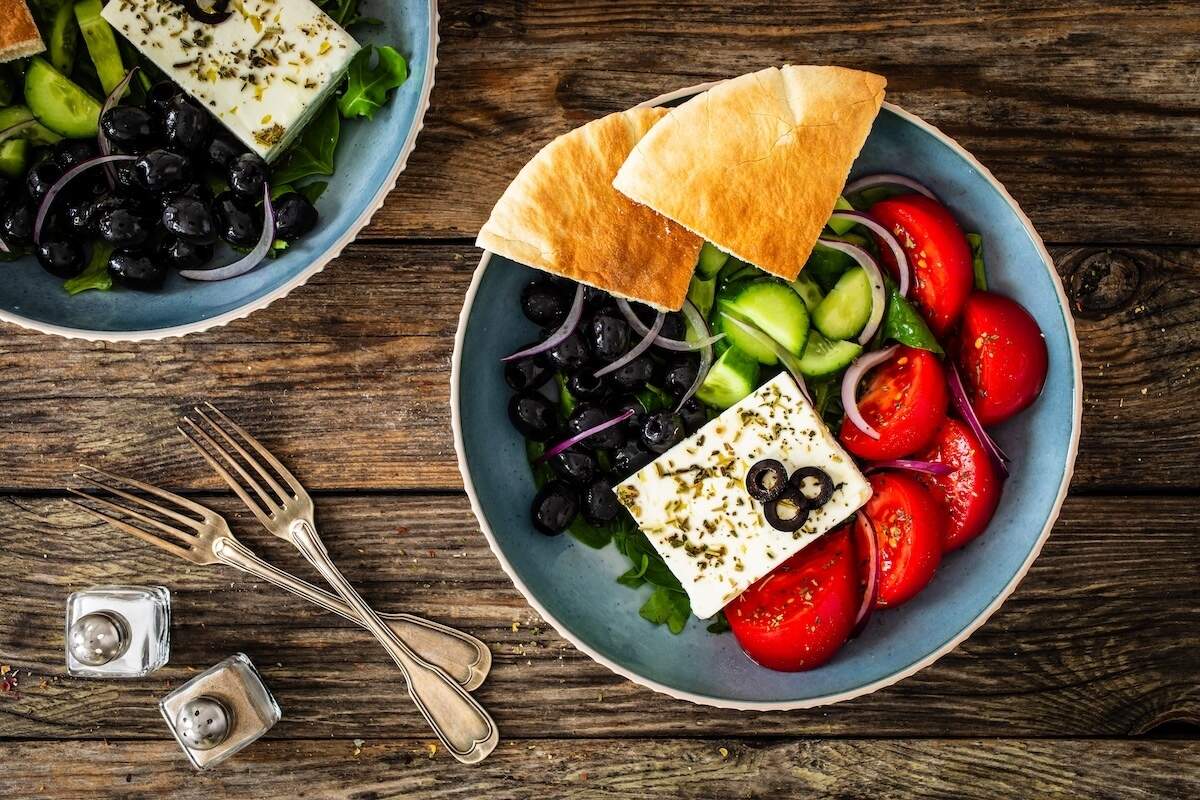 Top-down view of a Greek salad bowl on a rustic wooden table, featuring a block of feta cheese topped with herbs and olive slices, surrounded by sliced tomatoes, cucumbers, black olives, red onions, and two pieces of pita bread, with vintage forks and salt and pepper shakers on the side.