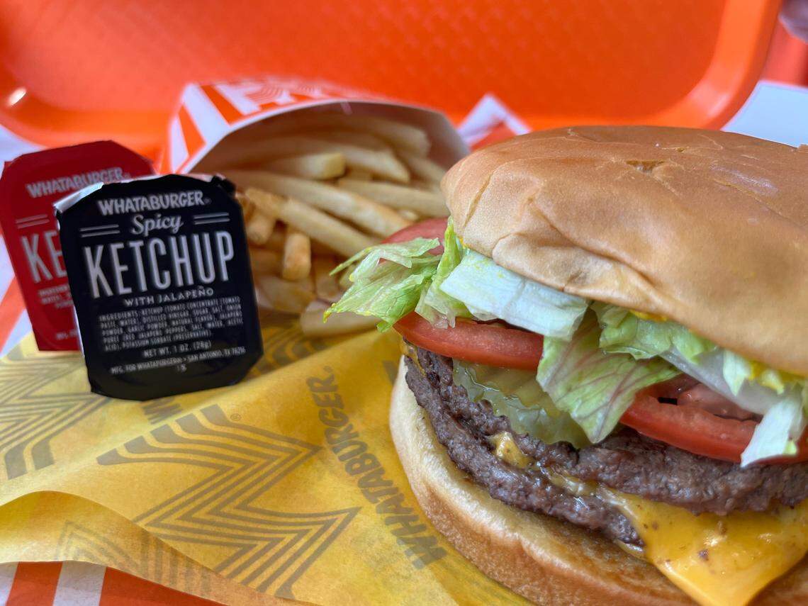A detailed shot of a Whataburger combo meal. In the foreground, a double cheeseburger with melted cheese, lettuce, and tomato sits on a yellow wrapper. Behind it are french fries and two tubs of ketchup, including the distinct black label for “Spicy Ketchup.” The meal is served on a bright orange tray.