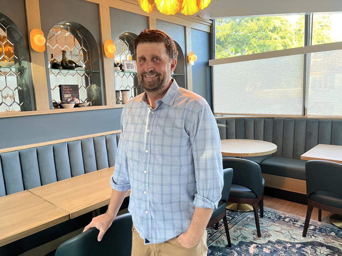 A friendly-looking restaurant owner with a beard stands in a private dining room while wearing a casual plaid button-down shirt. The restaurant interior is chic, with custom blue-grey booths, mirrored shelving and an elaborate amber-colored chandelier hanging from the ceiling.
