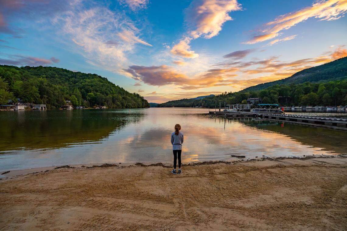 A young girl with her back to the camera stands alone on a sandy beach, looking out at a sunset over a calm lake. The vibrant orange, pink, and yellow clouds are reflected perfectly on the still water. The lake is surrounded by dark, densely forested hills, with boat docks and houses visible along the shoreline.