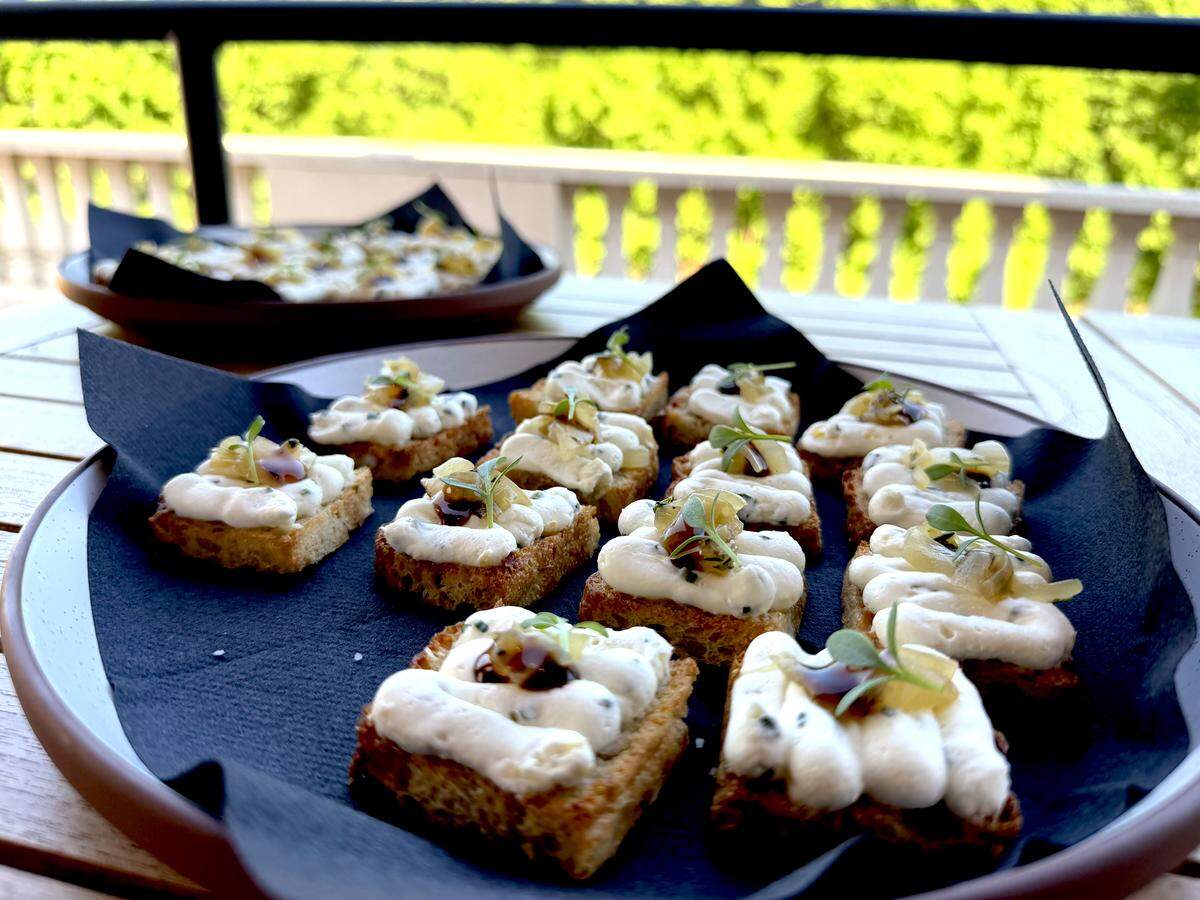 A close-up of a platter featuring small, square toasted bread slices topped with piped white cream or herbed goat cheese. Each appetizer is finished with a dark balsamic glaze, a small piece of preserved fruit, and a fresh microgreen garnish. The platter sits on a wooden outdoor table with a blurred green background.