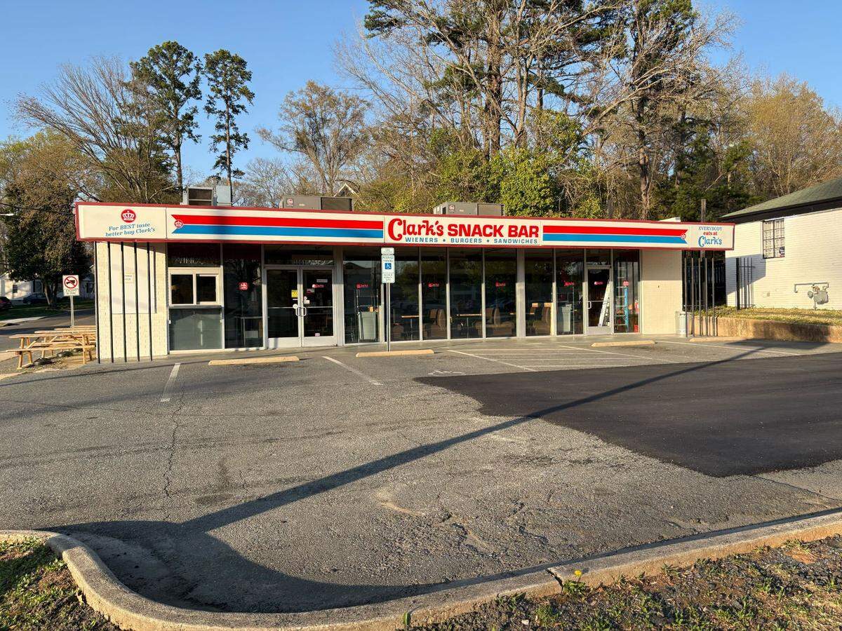 The exterior of Clark’s Snack Bar, a long, low-profile building with large glass windows and white brick walls. A retro-style sign runs across the top of the storefront with red and blue stripes and the text “Clark’s SNACK BAR” in bold red letters, followed by “WIENERS • BURGERS • SANDWICHES.” The restaurant faces an asphalt parking lot under a clear blue sky, with tall trees in the background and a few wooden picnic tables visible on the far left.