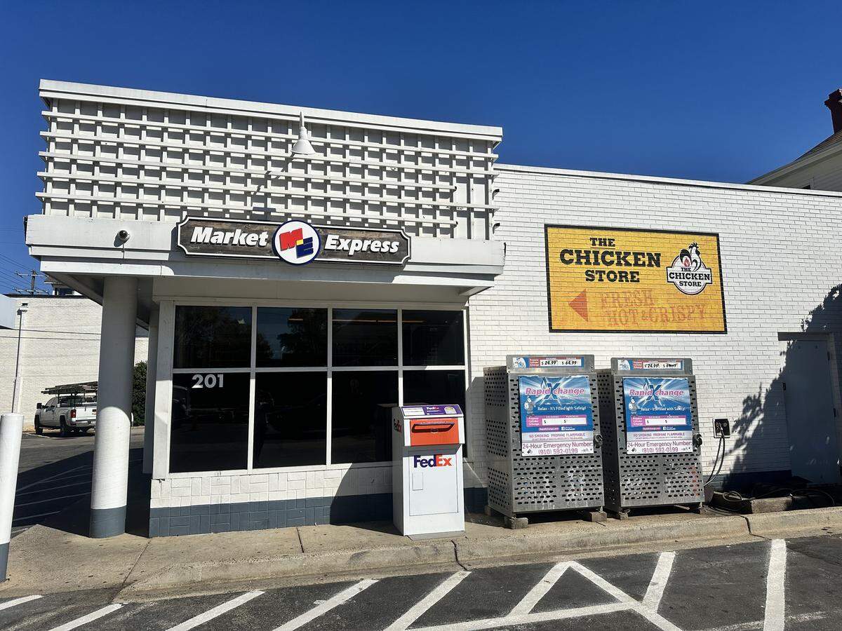 A straight-on, eye-level shot of the exterior of a “Market Express” convenience store on a sunny day. The one-story building is made of white painted brick and has a large, yellow, weathered sign for “THE CHICKEN STORE” on its right wall. In front of the store, on the sidewalk, are a FedEx drop box and two blue newspaper vending machines, all under a clear blue sky.