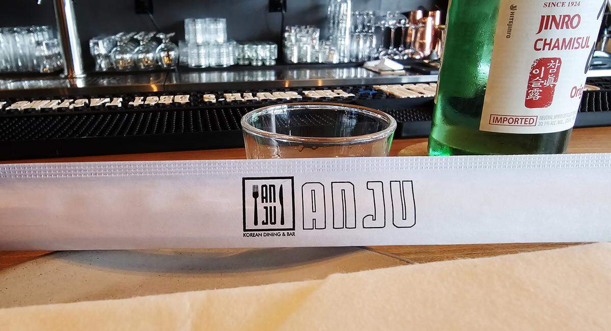 A close-up of a table setting at Anju, a Korean dining and bar. In the foreground, a paper chopstick sleeve features the restaurant’s logo. Next to it are a shot glass and a green bottle of Jinro Chamisul soju. The background shows the restaurant’s bar with neatly stacked glasses.