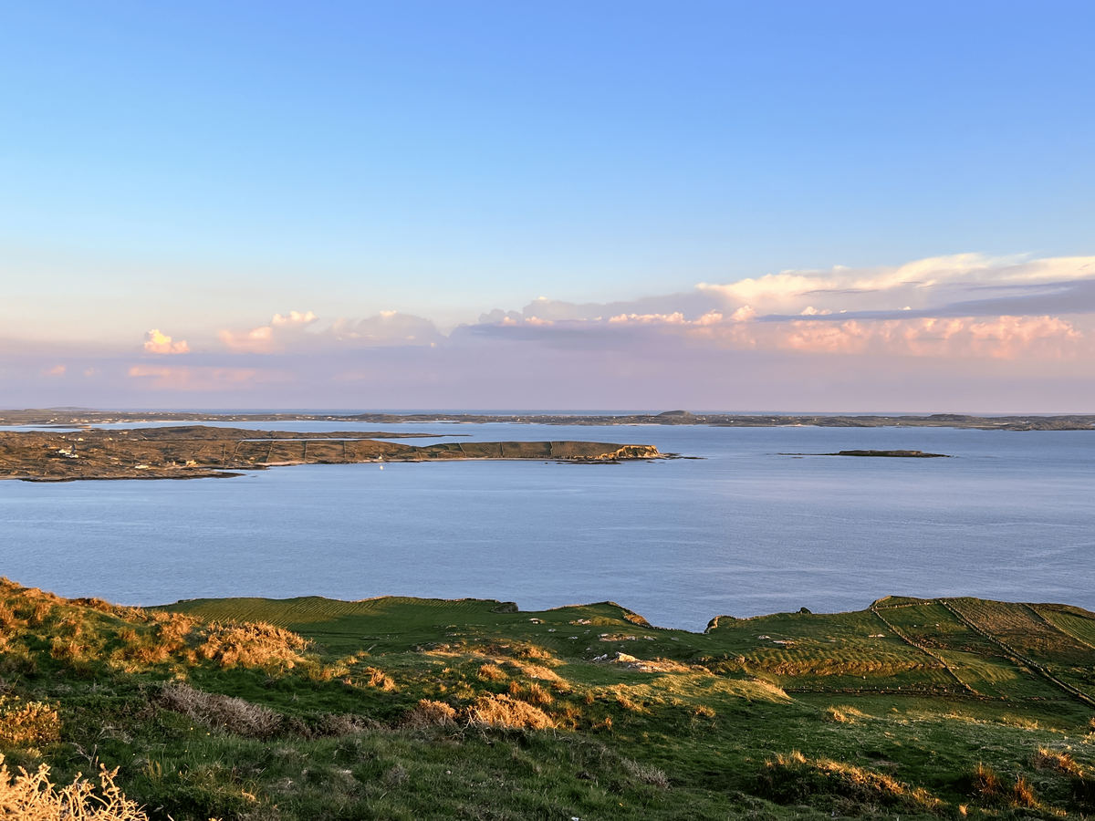 A breathtaking panoramic view of a calm sea or large bay at sunset. The sky is a gradient of light blue, pink, and orange, with fluffy white clouds reflecting the warm light. In the foreground, green hills and fields slope down toward the water. The sea is a serene blue, and several small islands or landmasses are visible on the horizon.