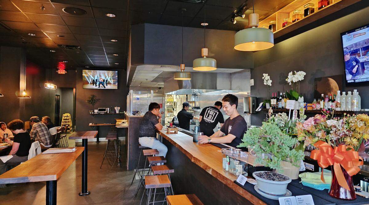 The warm and modern interior of a restaurant view shows a long wooden bar where a bartender is attending to customers seated on stools. Behind the bar, an open kitchen is bustling with activity. The space is decorated with a bonsai tree and a large flower arrangement, and other diners are seated at tables in the background.