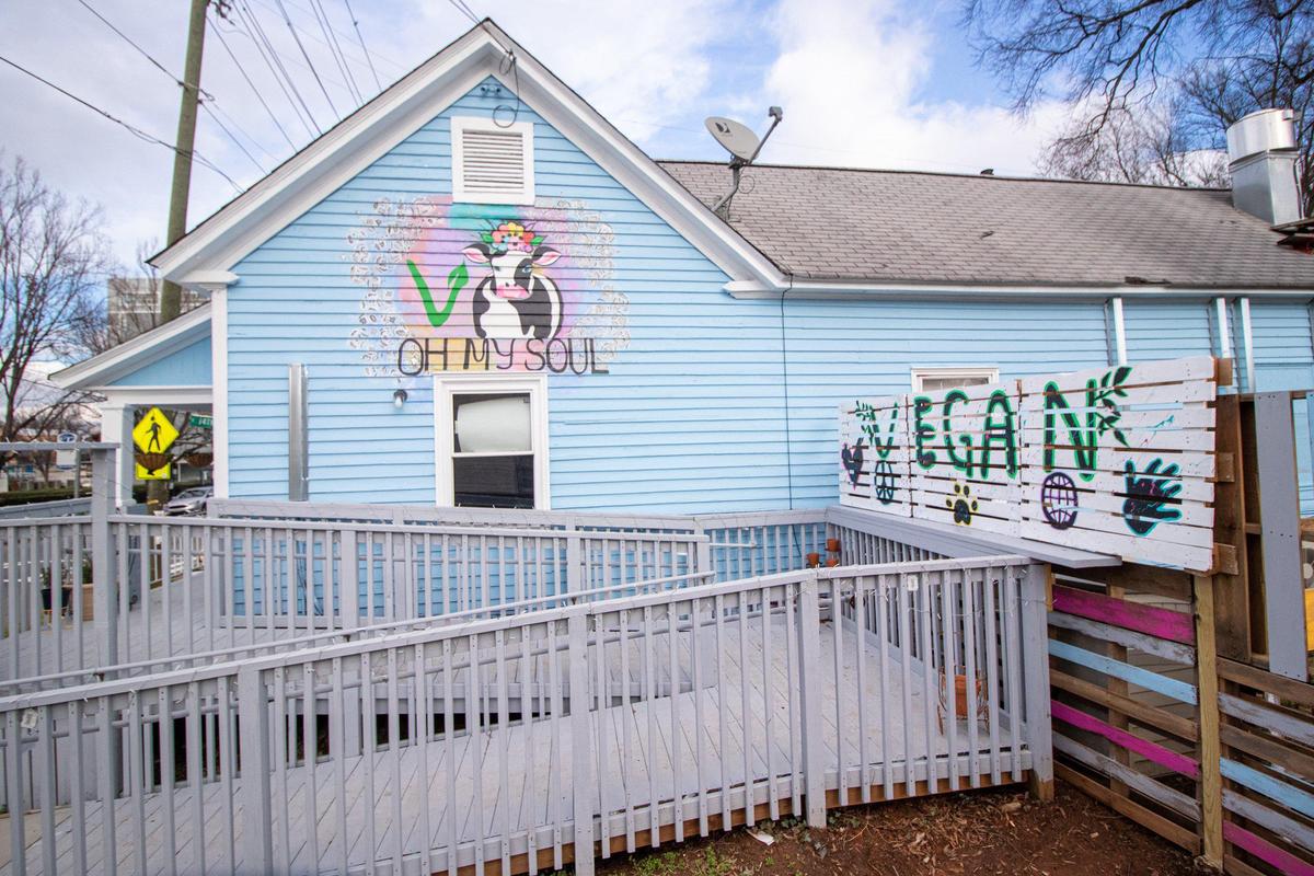 This is an exterior view of the Oh My Soul restaurant, which is painted a light blue with white trim. A colorful mural of a cow wearing sunglasses is painted above a window, and the entrance features a gray wooden ramp and patio area labeled with “VEGAN” art on a small fence.