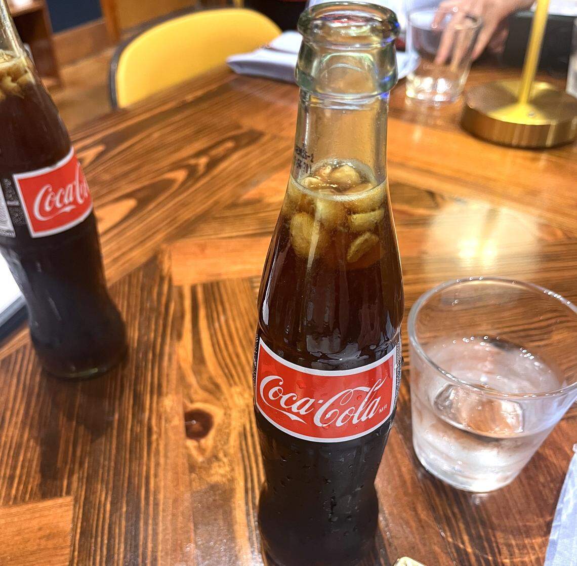 A detailed shot of a glass Coca-Cola bottle, beaded with condensation, on a restaurant table. Several ice cubes are visible inside the bottle’s opening. Another bottle and a glass of water are also on the rich, dark wood table.