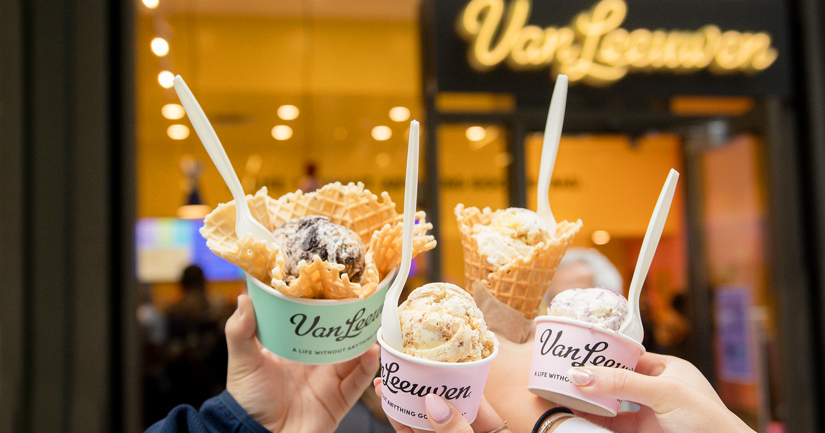 Several hands hold up four different ice cream treats—some in cups and some in waffle cones—in a “cheers” gesture. The treats are held in front of the blurred yellow storefront of a Van Leeuwen ice cream shop.