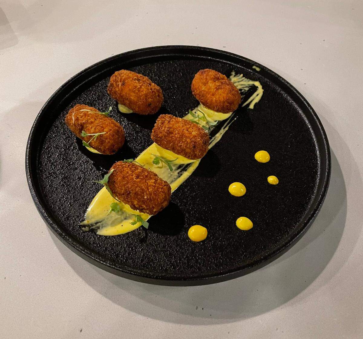 A dark, textured circular plate holds five golden-brown, oval-shaped croquettes, artfully arranged on a diagonal smear of light yellow sauce. Each croquette is garnished with a few green herbs. To the right of the main arrangement, several small, bright yellow dots of sauce are scattered on the plate. The plate rests on a light-colored, possibly white or light gray, countertop.