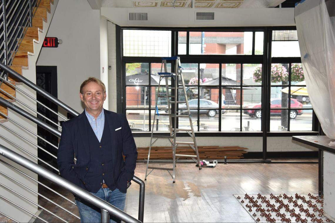 A smiling, middle-aged man wearing a dark blazer and blue jeans stands on a metal staircase inside a commercial space under construction. Behind him, large industrial windows look out onto a city street, and a ladder sits on the partially finished wood floor.
