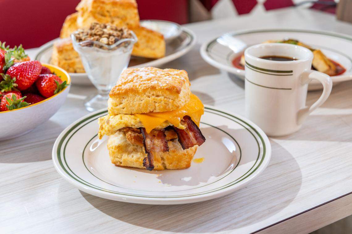 A high-angle shot of a breakfast table featuring a fried chicken biscuit sandwich with a folded egg, crispy bacon, and melted cheddar cheese. A mug of black coffee, a bowl of fresh strawberries, and a yogurt parfait are also visible on the light-colored wooden table.