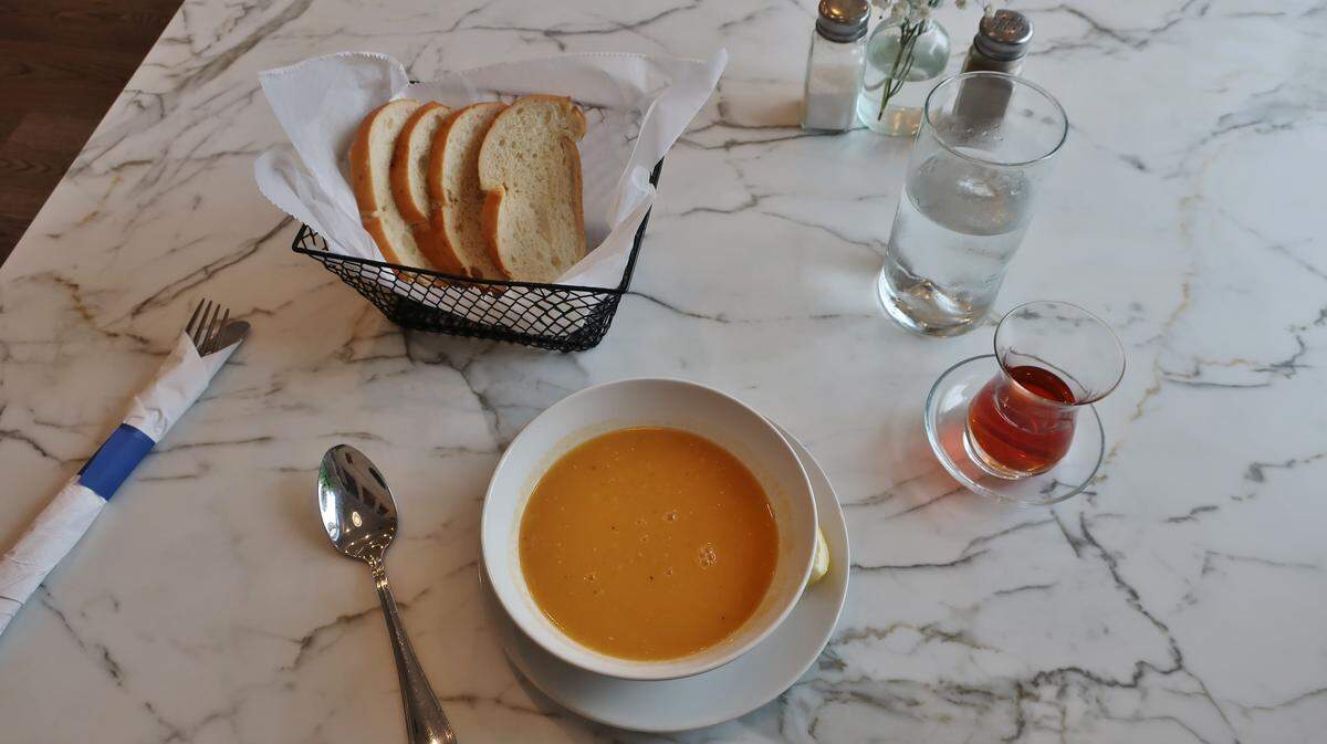 A bowl of Turkish lentil soup is served on a marble tabletop, accompanied by a basket of bread, a glass of water, and a traditional tulip-shaped glass of Turkish tea.