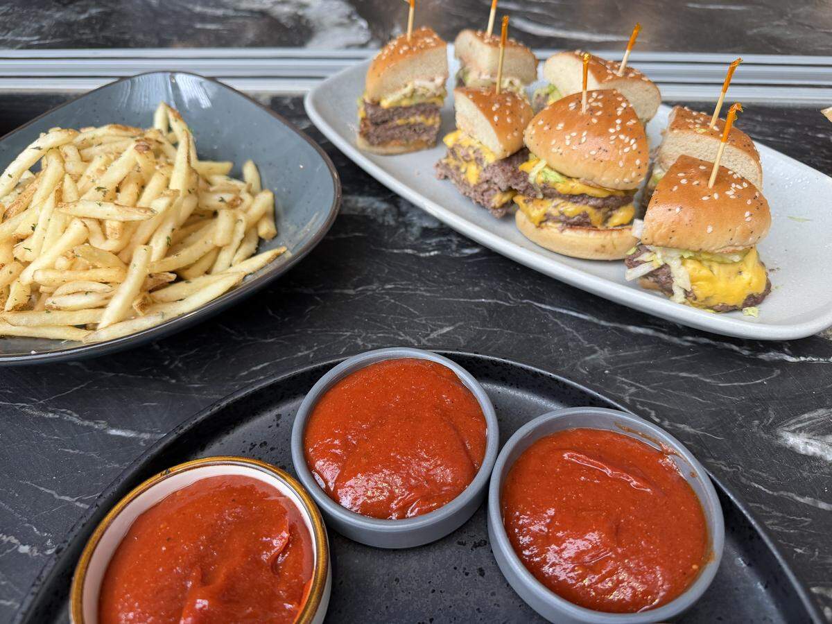 A slightly high-angle shot of a table spread featuring sliders, fries, and dipping sauces on a dark marbled surface. In the foreground, three small round dishes are filled with bright red sauce. Behind them, a gray ceramic bowl holds a large pile of thin, seasoned french fries, while a long white platter displays several small cheeseburger sliders on sesame seed buns, each secured with a wooden toothpick.