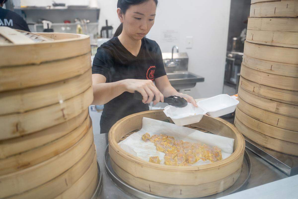 Surrounded by towering stacks of bamboo steamer baskets, a food stall employee in a black shirt carefully uses tongs to place bite-sized yellow dumplings into a takeout container. Steam rises from the open steamer in front of her.