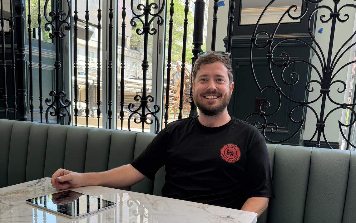 A restaurateur sits at a white marble table inside a restaurant with upscale decor, featuring teal banquette seating and ornate black wrought-iron partitions.