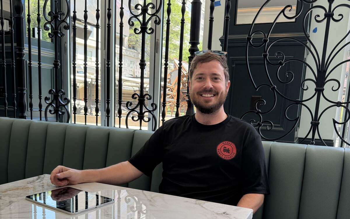 A restaurateur sits at a white marble table inside a restaurant with upscale decor, featuring teal banquette seating and ornate black wrought-iron partitions.