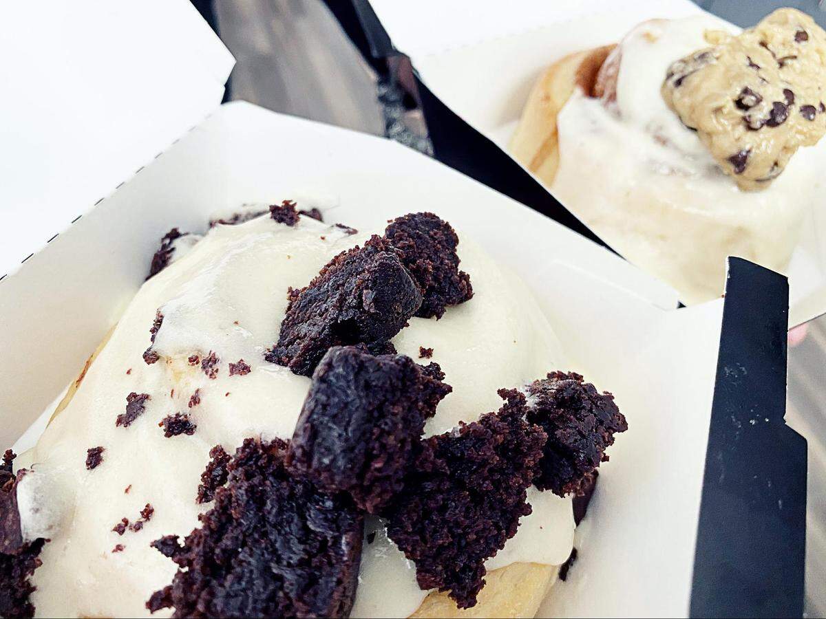 Cinnaholic cinnamon rolls with peanut butter frosting topped with brownie bites and one with cream cheese frosting topped with cookie dough.