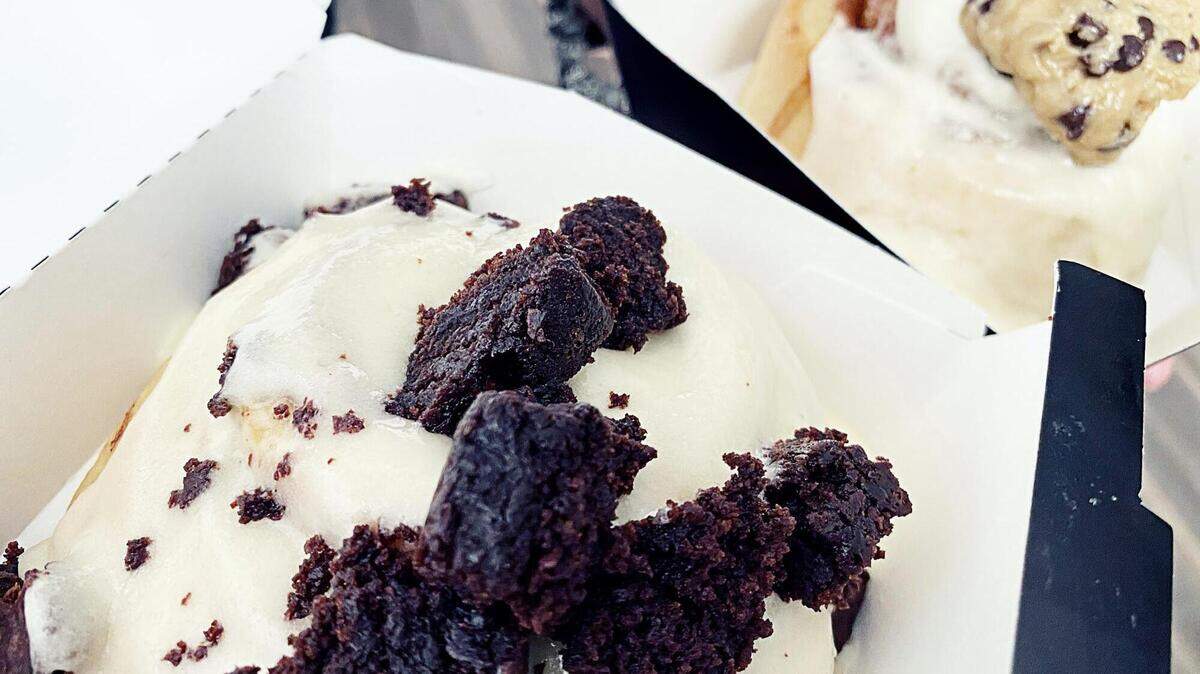 Cinnaholic cinnamon rolls with peanut butter frosting topped with brownie bites and one with cream cheese frosting topped with cookie dough.