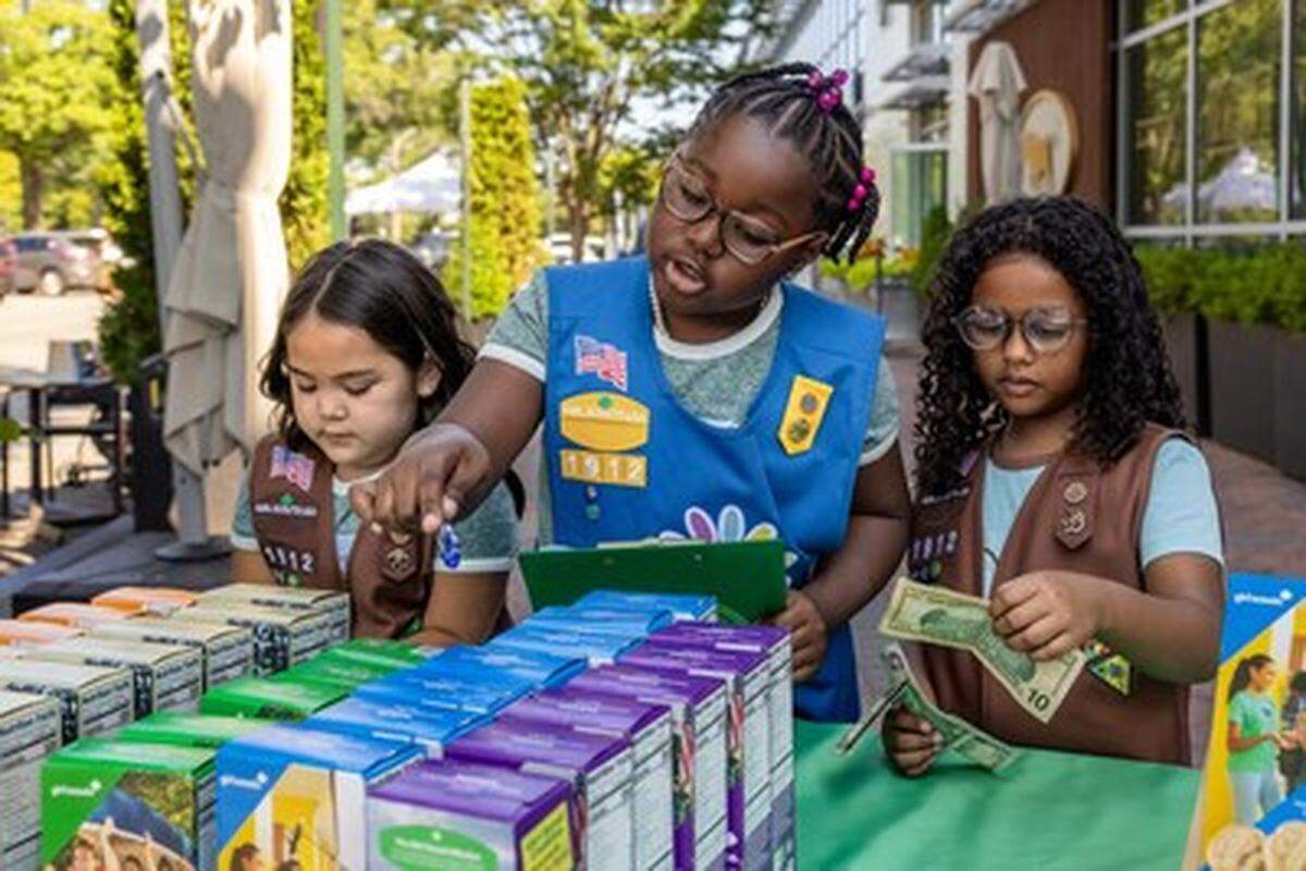 Three Girl Scouts stand behind a table filled with colorful cookie boxes at an outdoor booth. One scout manages a clipboard while another counts paper currency, demonstrating hands-on business and money management skills.