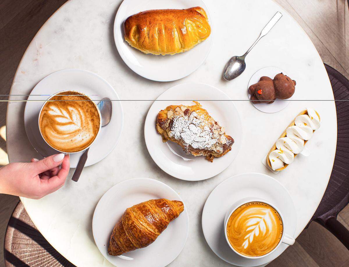 An overhead, flat-lay photograph of a marble cafe table laden with an assortment of gourmet pastries and two lattes. In the upper-left and middle-right sections of the frame are two white cups of coffee featuring intricate white leaf-patterned latte art. A person’s hand is visible on the right, gently touching the rim of one cup.