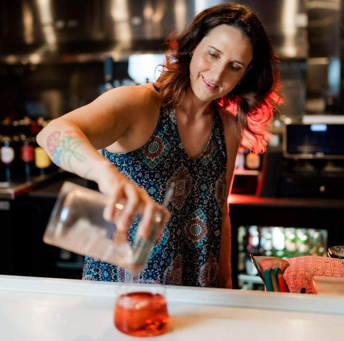 A bartender with wavy, shoulder-length brown hair leaning over a bright white bar counter to pour a drink while wearing a dark blue patterned halter top and showing of a floral tattoo on her forearm. The bartender is pouring a clear liquid from a mixing glass into a short glass containing a red cocktail and a large ice cube.