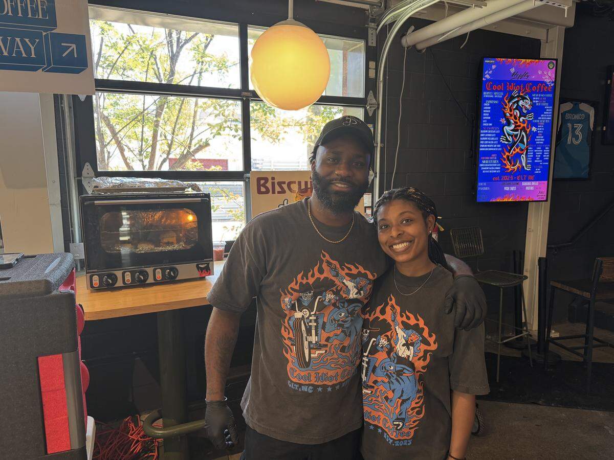 A man and a woman stand side-by-side inside a coffee shop, smiling warmly at the camera. They are both wearing matching black t-shirts with a vibrant orange and blue graphic of a character on a motorcycle, which reads “Cool Idiot Coffee.” In the background, a small countertop oven contains warming biscuits, and a digital menu board displays specialty drink options.