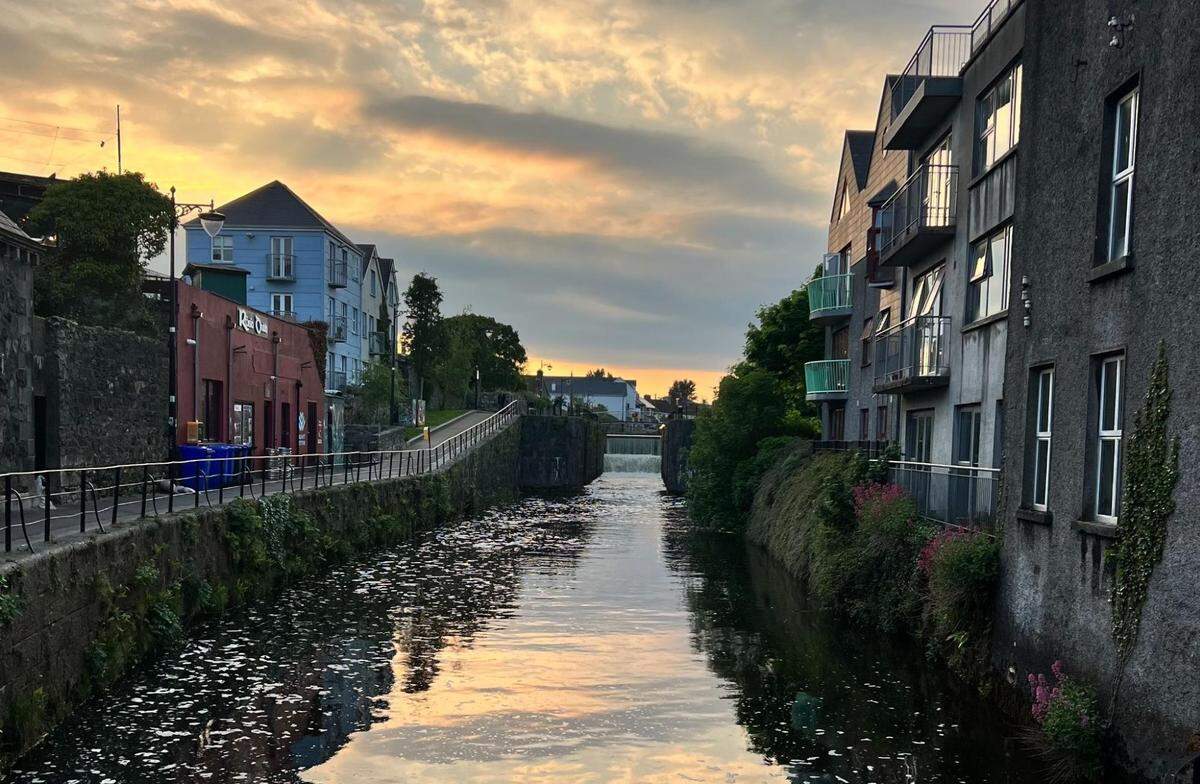 A picturesque view of a narrow river flowing between two rows of buildings. The sky is a beautiful blend of orange and pink from the sunset, which is reflected in the calm water. The buildings on the left are a mix of colorful facades, and the buildings on the right are a modern gray with balconies. There is a small waterfall or dam in the distance, and the river is lined with a low stone and metal wall.
