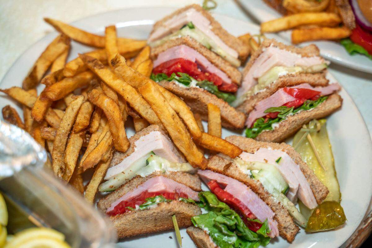 A restaurant plate is generously filled with a quartered club sandwich and a large side of french fries. The sandwich’s layers of sliced turkey, ham, tomato, and greens are visible. The fries are golden and appear crispy.