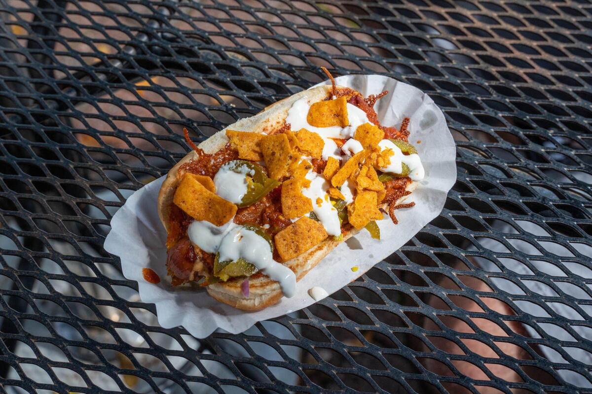 A top-down, close-up shot of a hot dog is served in a paper tray. The hot dog is piled high with a dark red chili, what looks like diced onions, and green jalapeño slices. It is generously topped with a dollop of sour cream or a white sauce and sprinkled with crushed orange corn chips. The hot dog is nestled in a split bun and rests on a black metal mesh table.