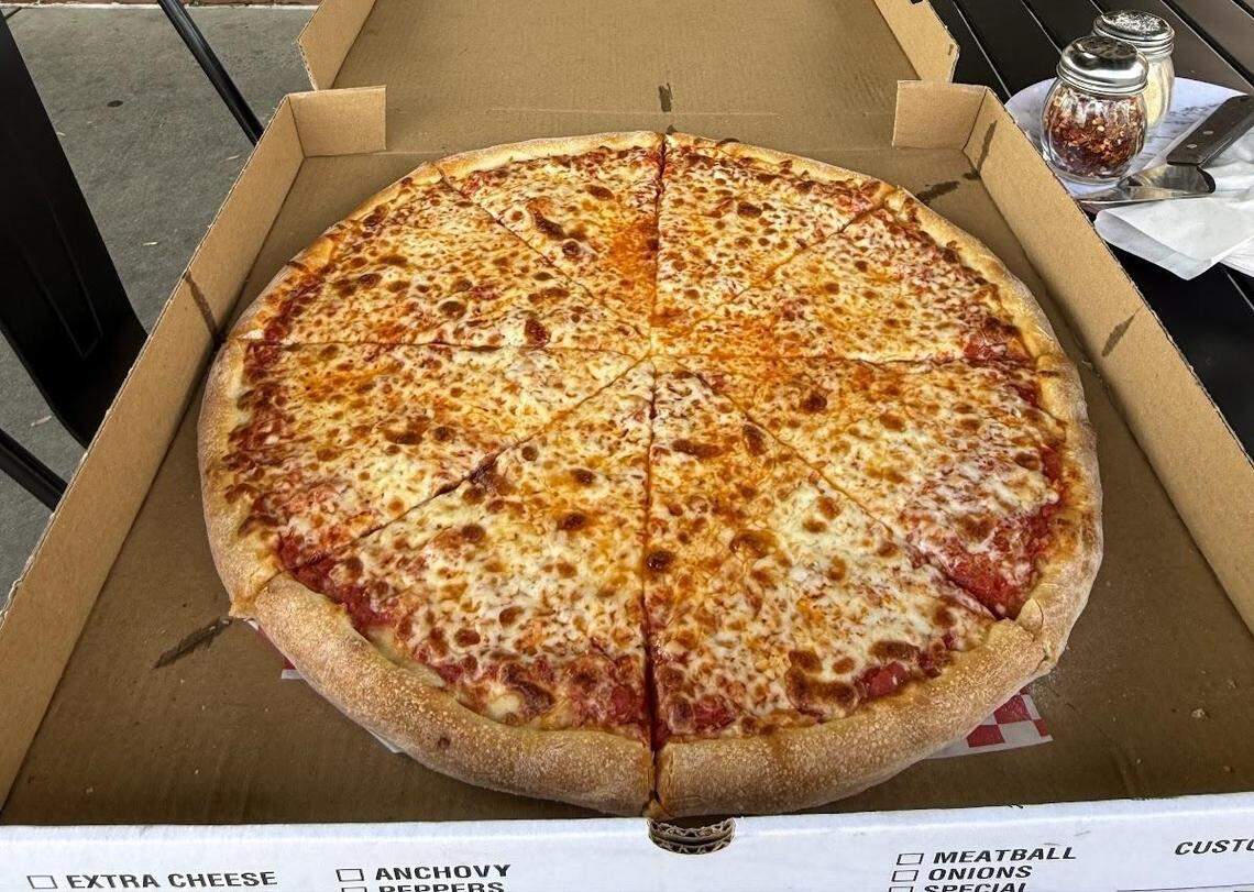 A top-down shot of a large, round cheese pizza in a cardboard takeout box. The pizza is cut into eight slices and has a bubbly, golden-brown cheese topping with a visible layer of red sauce. The crust is thick and golden. The edge of a red and white checkered paper liner is visible. The pizza box rests on a dark outdoor table, with shakers for red pepper flakes and grated cheese, along with a knife and fork, in the upper right corner.
